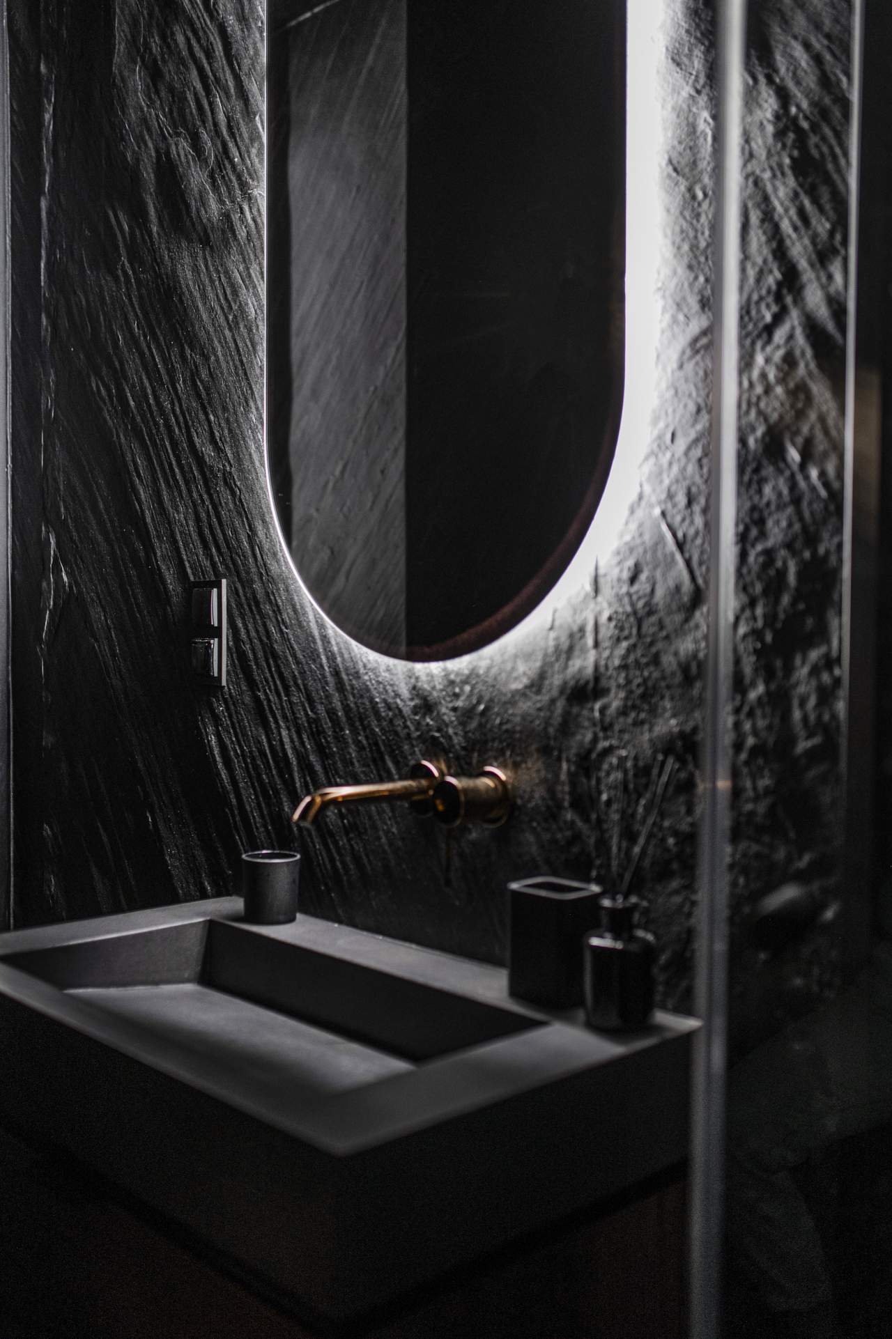 Masowa Stone - Stone Veneer Experts, UK-wide from Arbroath. Modern dark bathroom sink with a wall-mounted faucet and an illuminated oval mirror on textured wall.