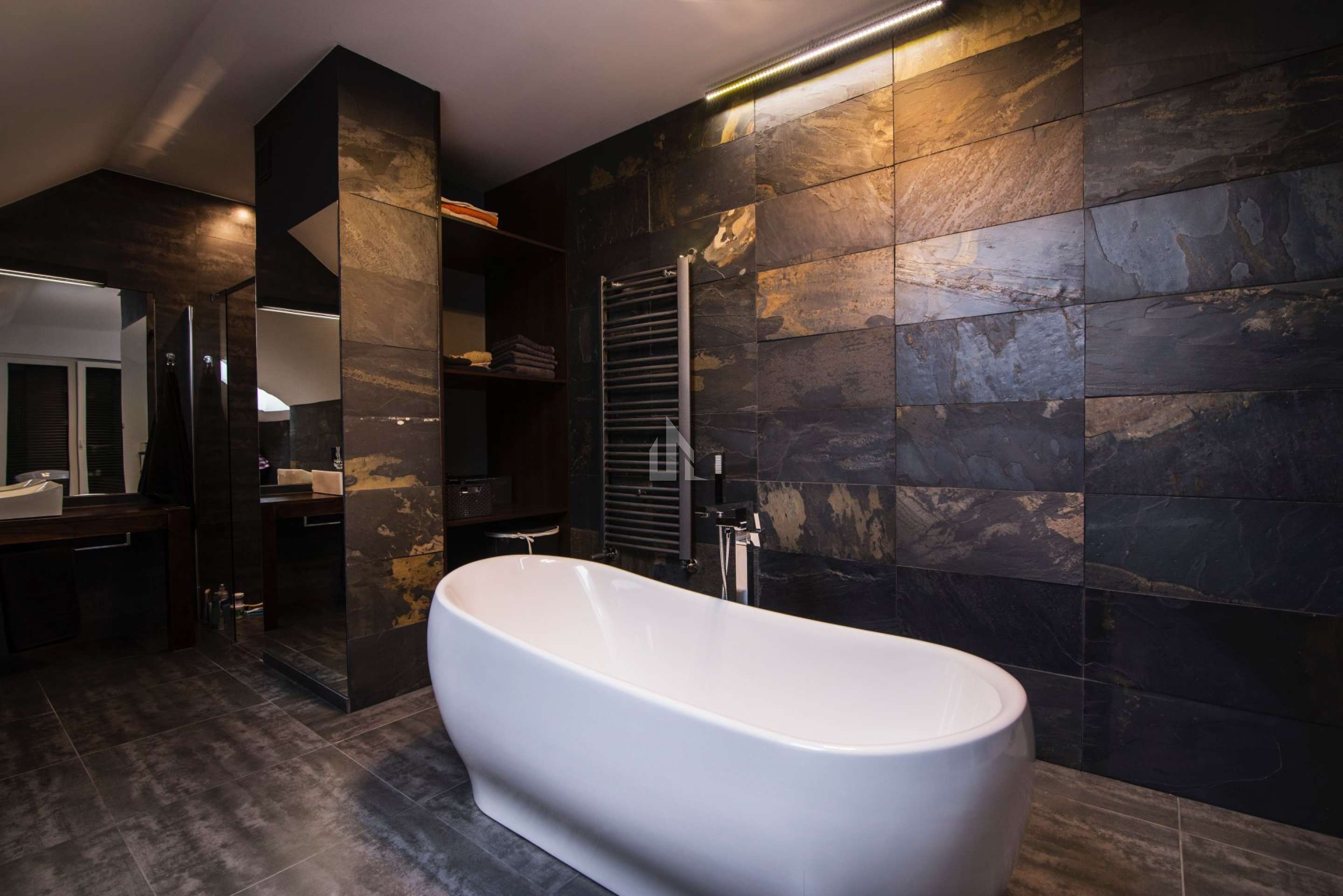 Masowa Stone - Stone Veneer Experts, UK-wide from Arbroath. Modern bathroom with a freestanding white bathtub, dark stone tiled walls and floor, and shelves with folded towels.